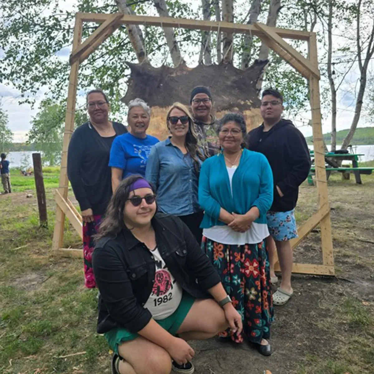 Groupe de personnes réunies à l’extérieur sous une structure en bois, souriant et posant ensemble